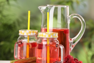 Mason jars and jug of tasty rhubarb cocktail with raspberry outdoors, closeup