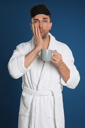 Sleepy young man in bathrobe with cup of coffee on blue background