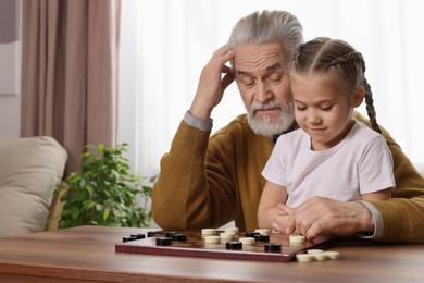 Photo of Playing checkers. Grandfather learning little girl at table in room
