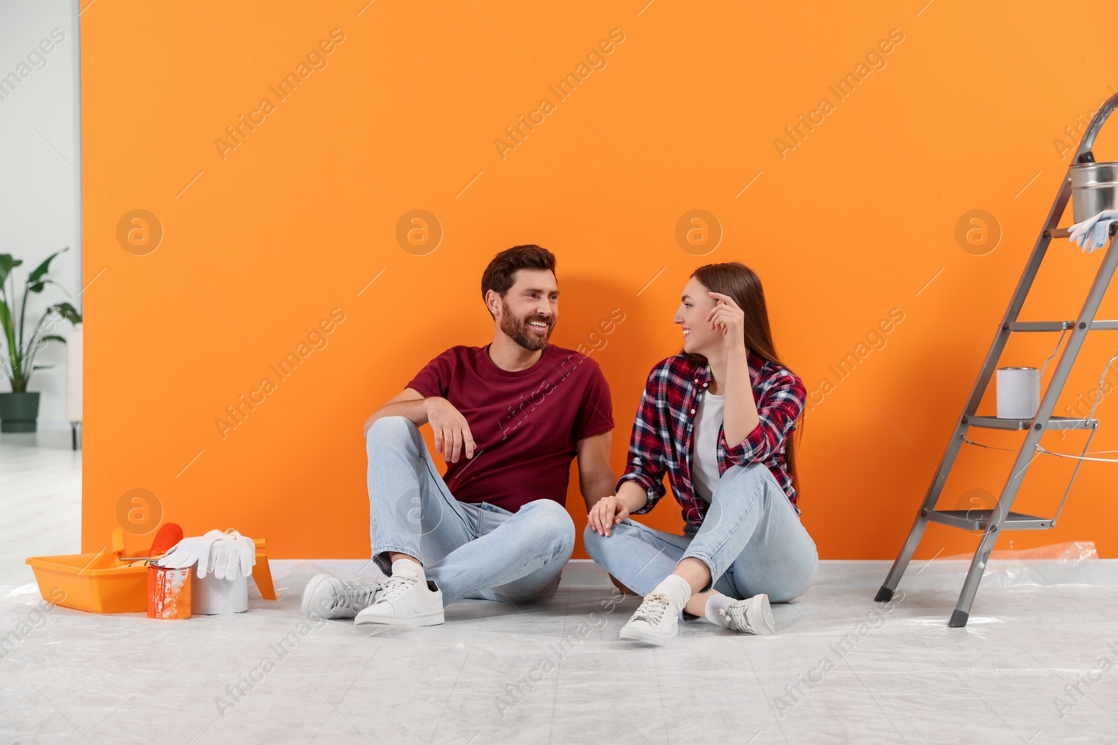 Photo of Happy designers sitting on floor with painting equipment near freshly painted orange wall indoors