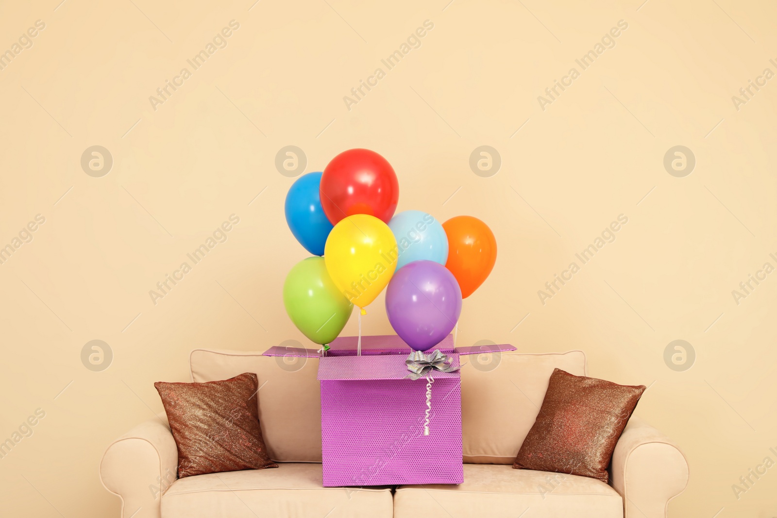 Photo of Gift box with bright air balloons on sofa against color background