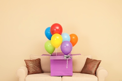 Gift box with bright air balloons on sofa against color background