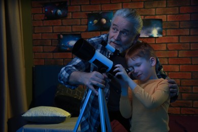 Photo of Little boy with his grandfather looking at stars through telescope in room