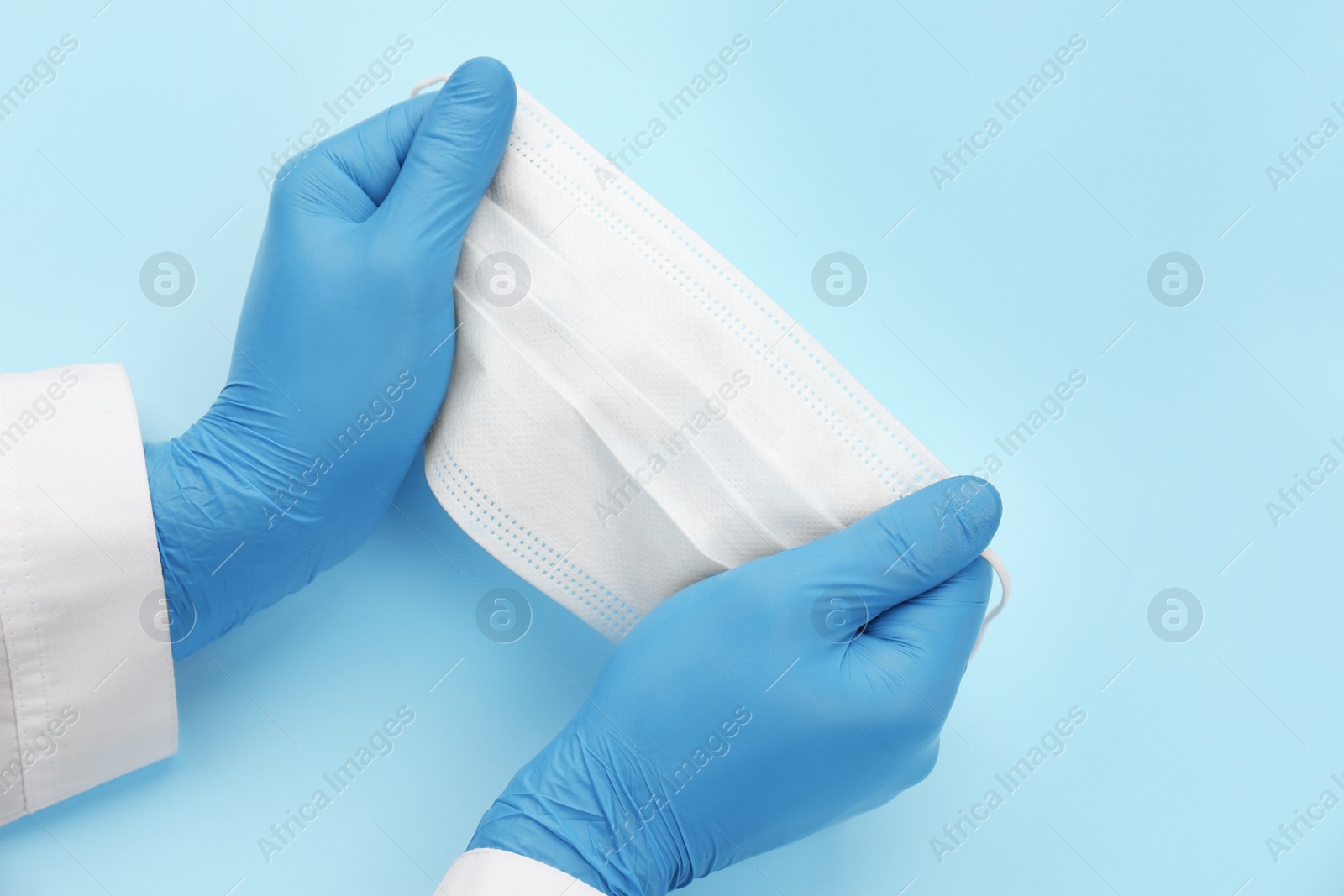 Photo of Doctor in medical gloves holding protective mask on light blue background, closeup. Space for text