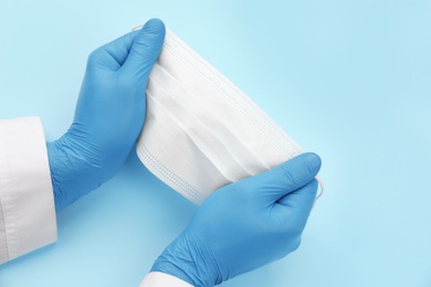 Doctor in medical gloves holding protective mask on light blue background, closeup. Space for text