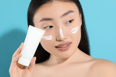 Beautiful young woman with sunscreen on her face holding sun protection cream against light blue background, closeup