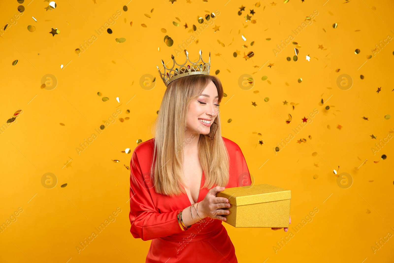 Photo of Happy young woman in party crown with gift box and confetti on yellow background