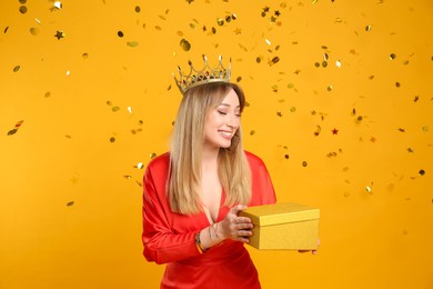 Happy young woman in party crown with gift box and confetti on yellow background