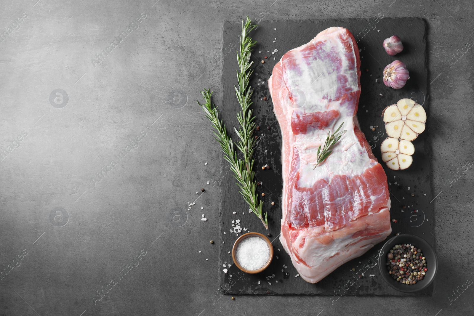 Photo of Piece of raw pork belly, rosemary, garlic and spices on grey textured table, top view. Space for text