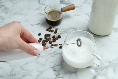 Woman whisking milk in cup with mini mixer (milk frother) at white marble table, closeup