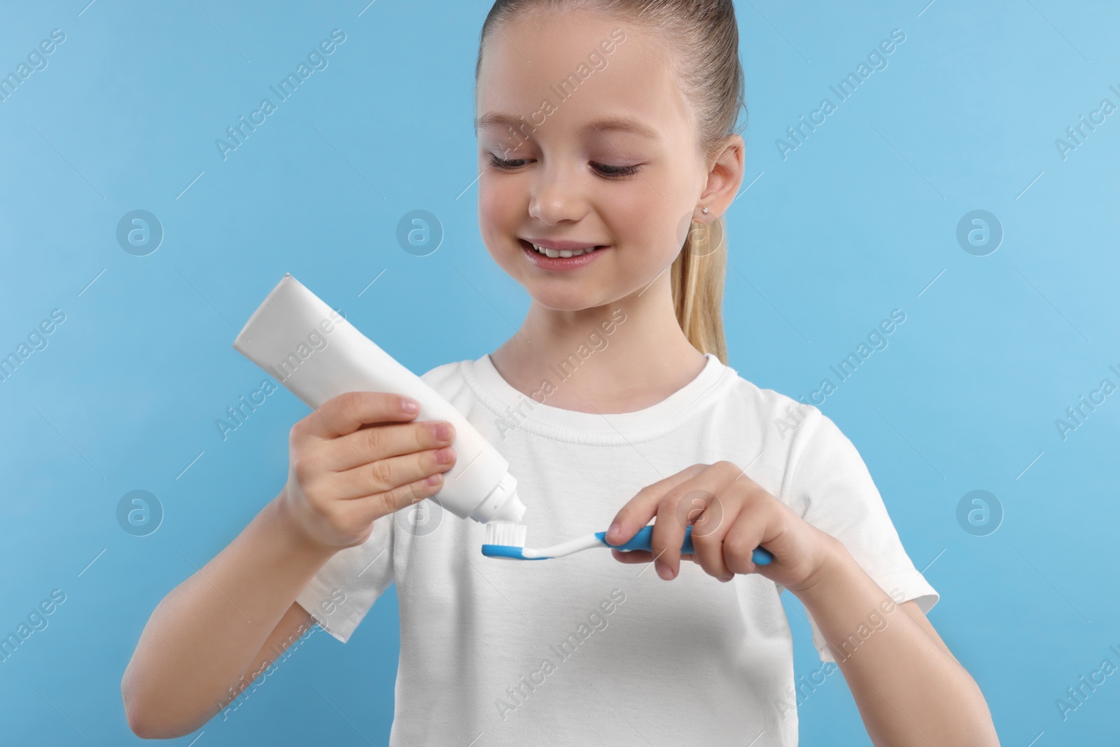 Photo of Happy girl squeezing toothpaste from tube onto toothbrush on light blue background