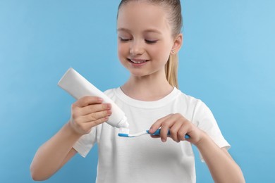 Photo of Happy girl squeezing toothpaste from tube onto toothbrush on light blue background
