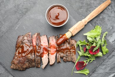 Photo of Pieces of delicious roasted beef meat with sauce and greens on black table, top view