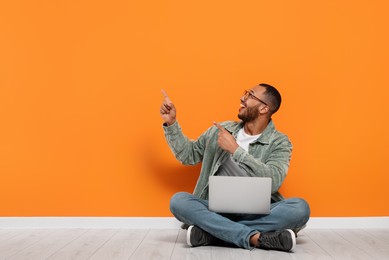 Smiling young man with laptop sitting on floor near orange wall, space for text
