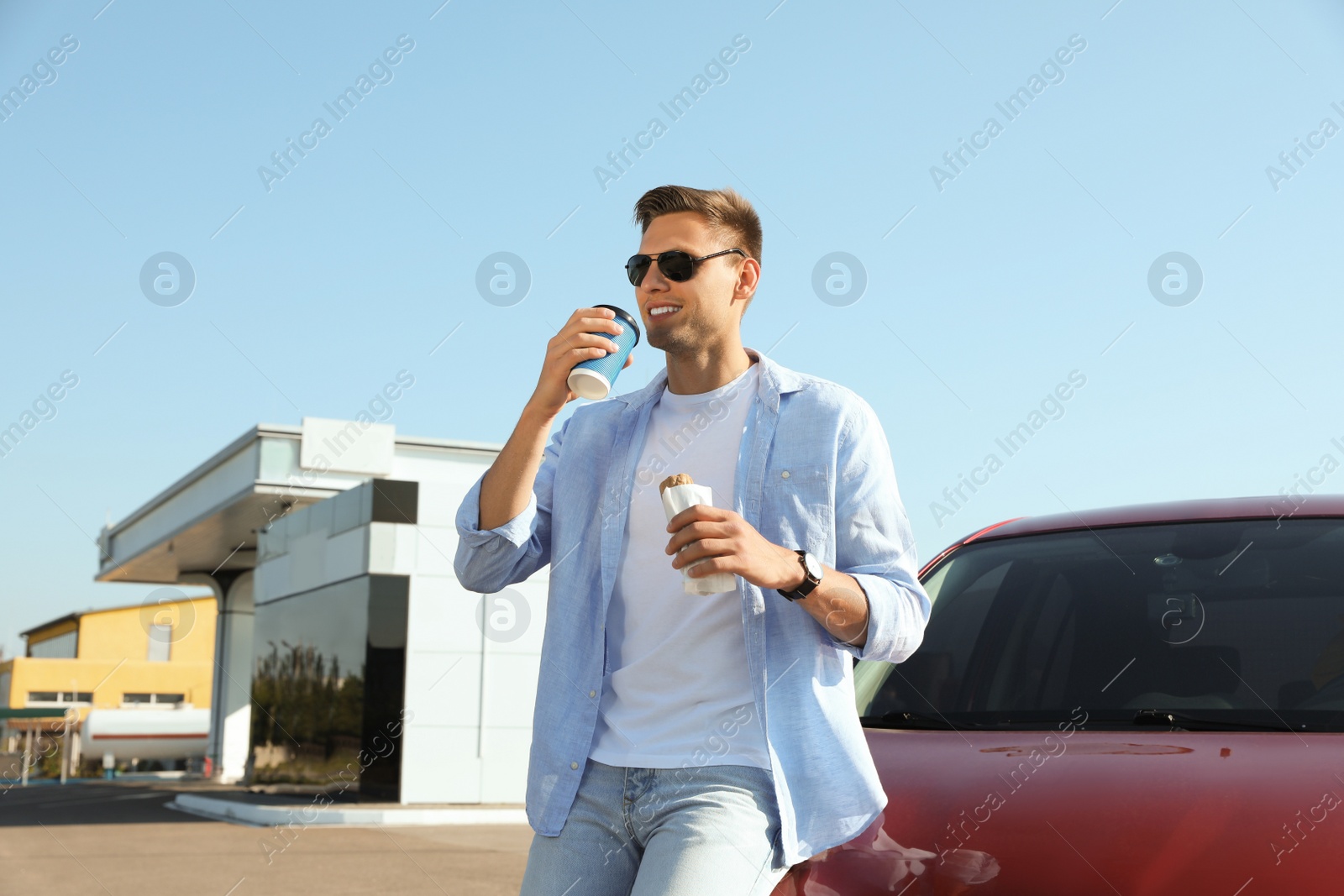 Photo of Young man with hot dog drinking coffee near car at gas station