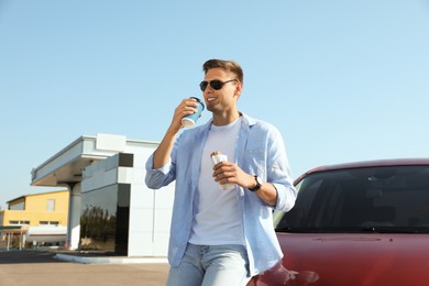 Photo of Young man with hot dog drinking coffee near car at gas station