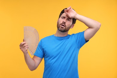 Unhappy man with hand fan suffering from heat on orange background