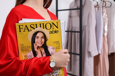 Young woman with fashion magazine near clothes rack indoors, closeup
