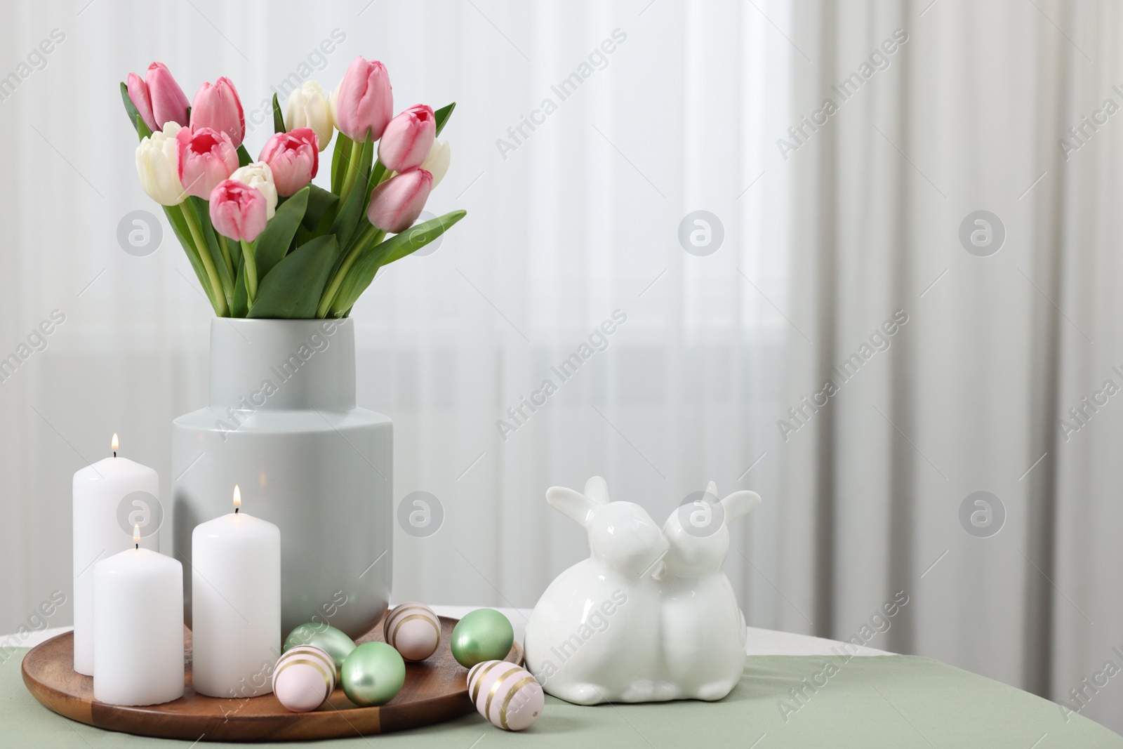 Photo of Easter decorations. Bouquet of tulips, painted eggs, burning candles and bunny figures on table indoors. Space for text