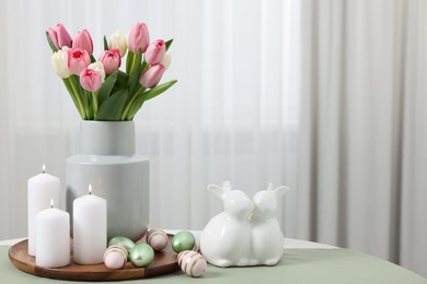 Easter decorations. Bouquet of tulips, painted eggs, burning candles and bunny figures on table indoors. Space for text