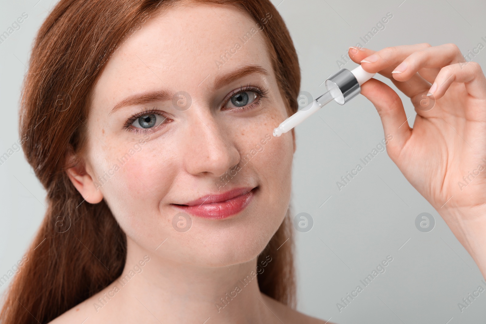 Photo of Beautiful woman with freckles applying cosmetic serum onto her face against grey background, closeup