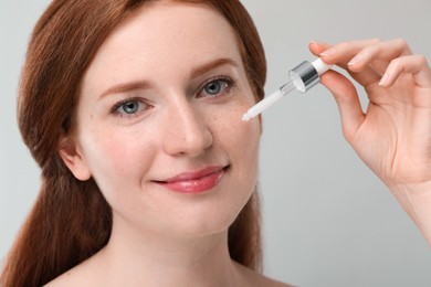 Beautiful woman with freckles applying cosmetic serum onto her face against grey background, closeup