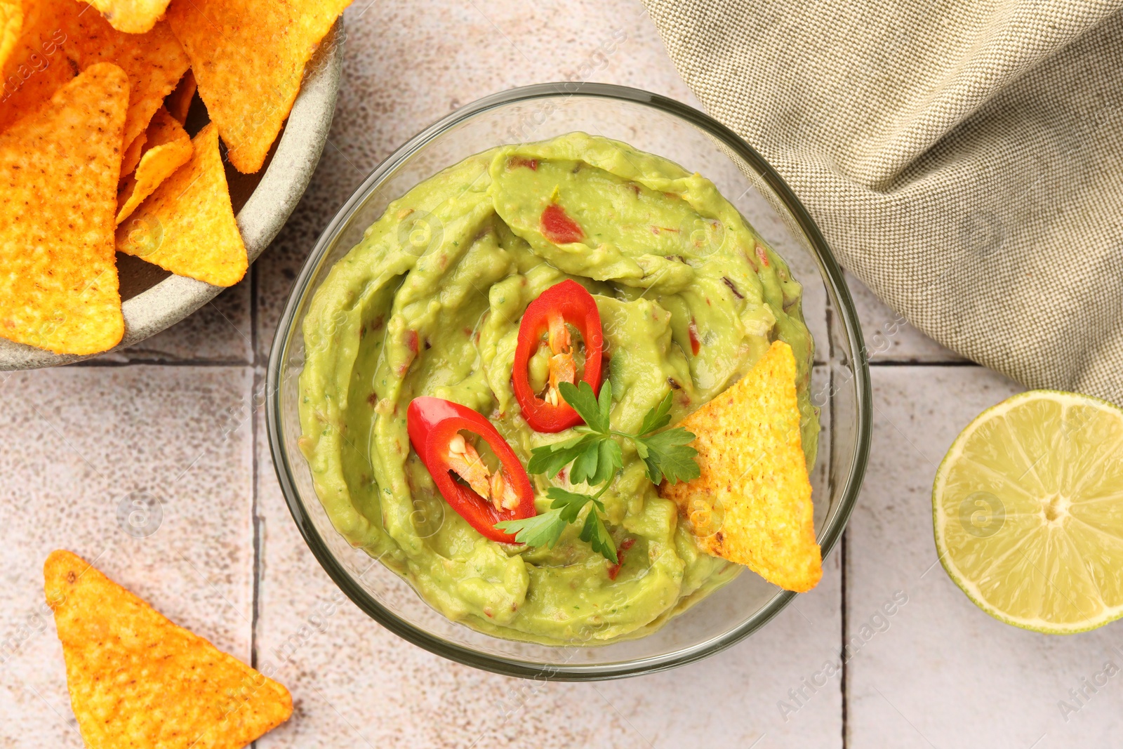 Photo of Bowl of delicious guacamole with chili pepper, nachos chips and lime on white tiled table, flat lay