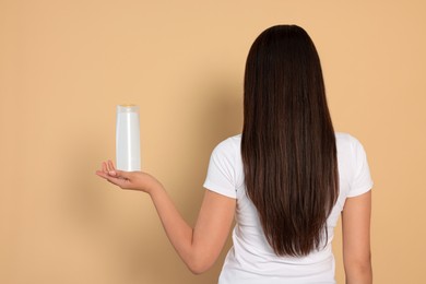 Young woman holding bottle of shampoo on beige background, back view
