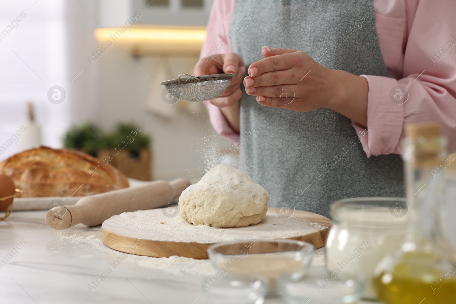 Photo of Making bread. Woman sifting flour onto dough at white table in kitchen, closeup