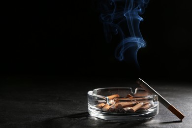 Photo of Glass ashtray with stubs and smoldering cigarette on grey table against black background. Space for text