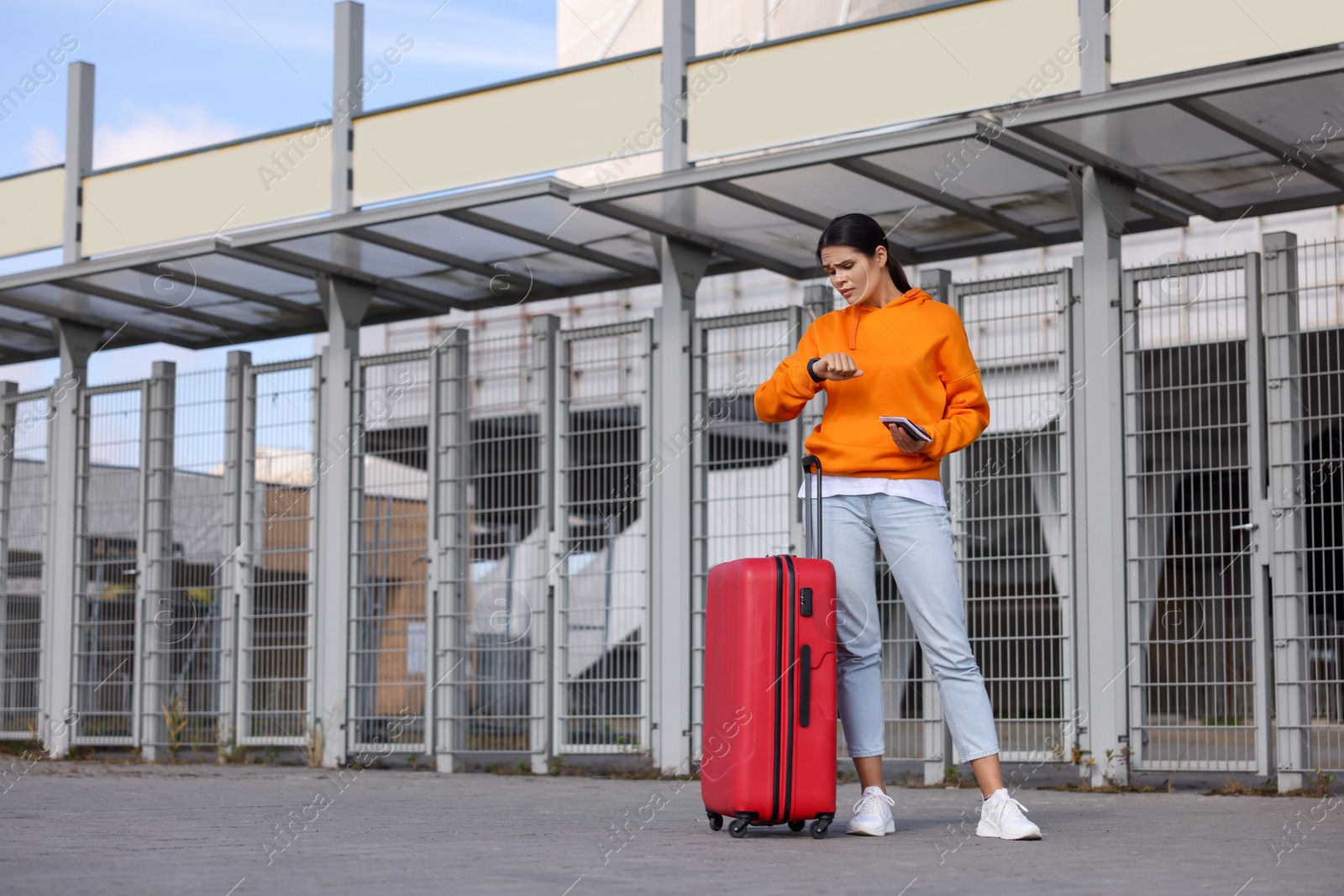 Photo of Being late. Worried young woman with red suitcase and passport looking at her watch outdoors, space for text