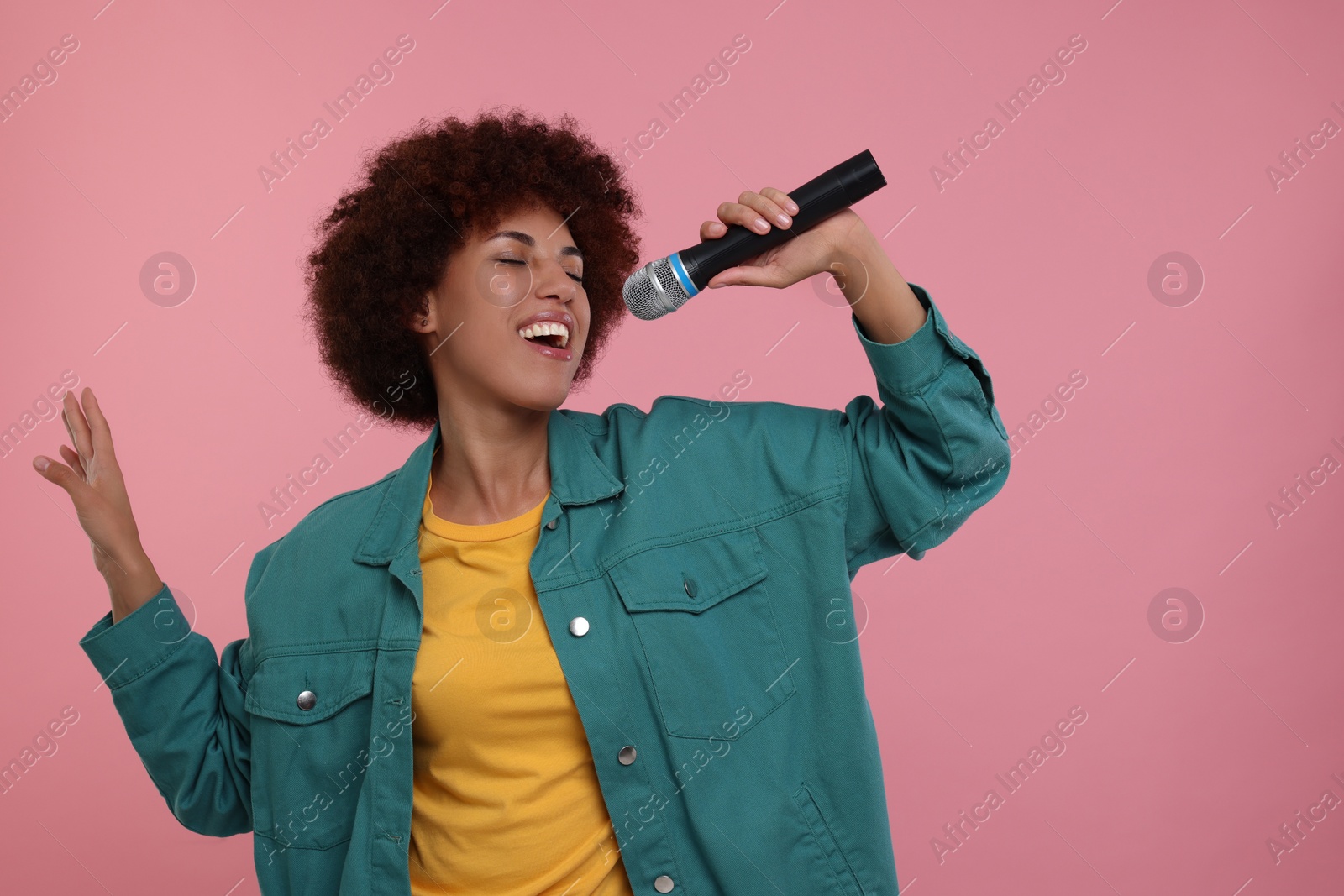 Photo of Curly young woman with microphone singing on pink background, space for text