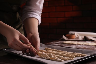 Woman putting homemade breadsticks on baking sheet at wooden table indoors, closeup. Cooking traditional grissini