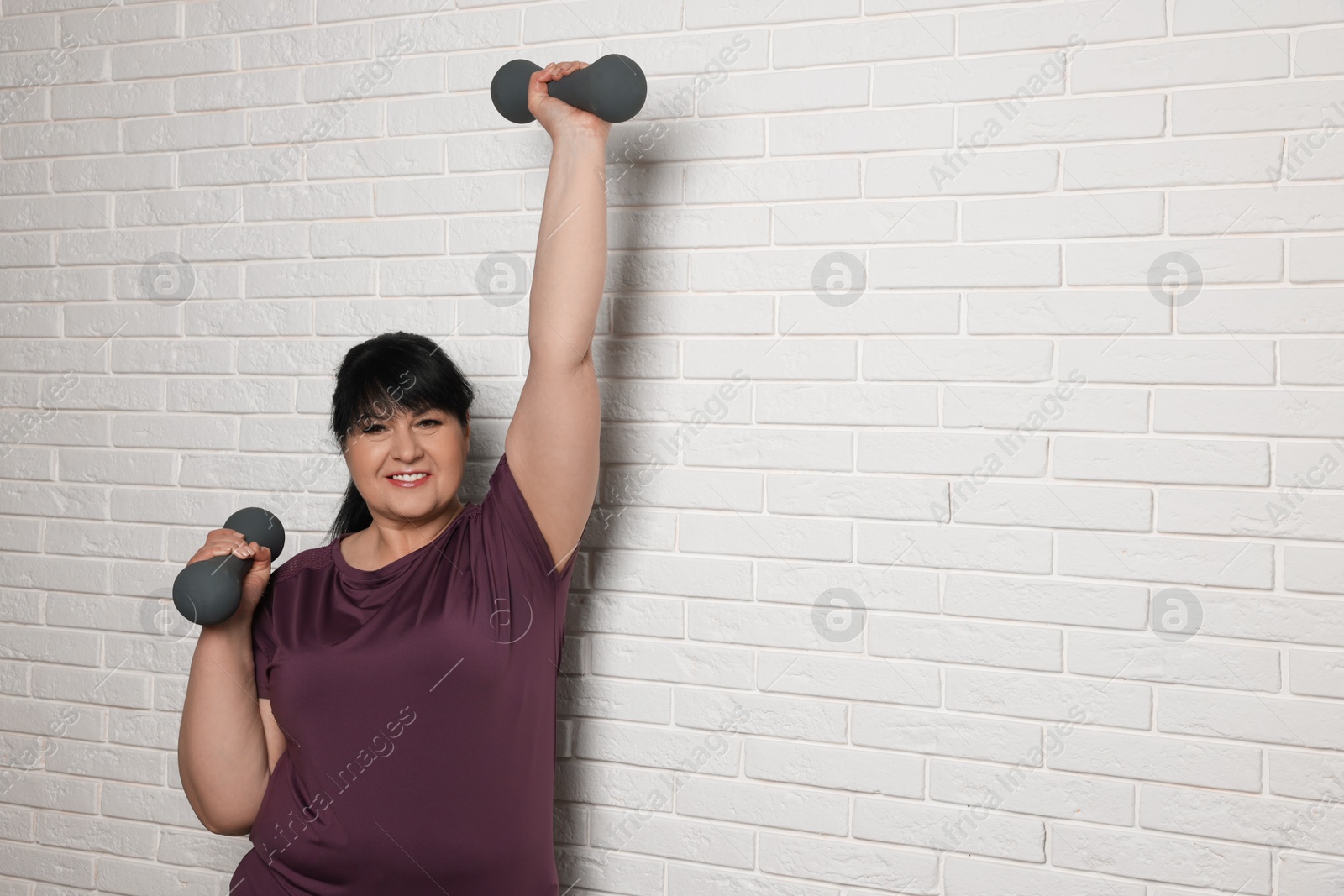 Photo of Happy overweight mature woman doing exercise with dumbbells near white brick wall, space for text