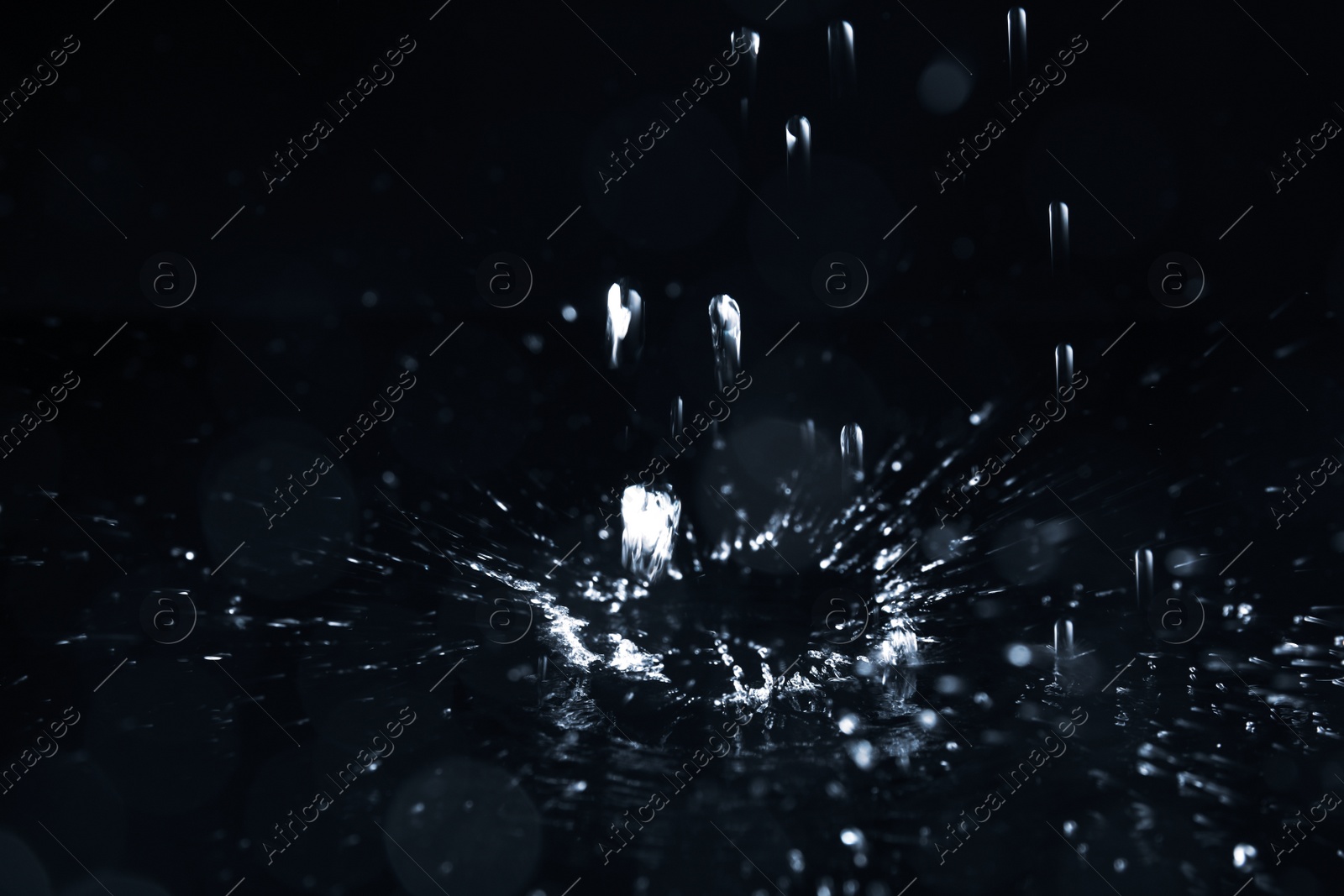 Photo of Rain drops falling down into puddle on dark background, closeup