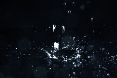 Photo of Rain drops falling down into puddle on dark background, closeup