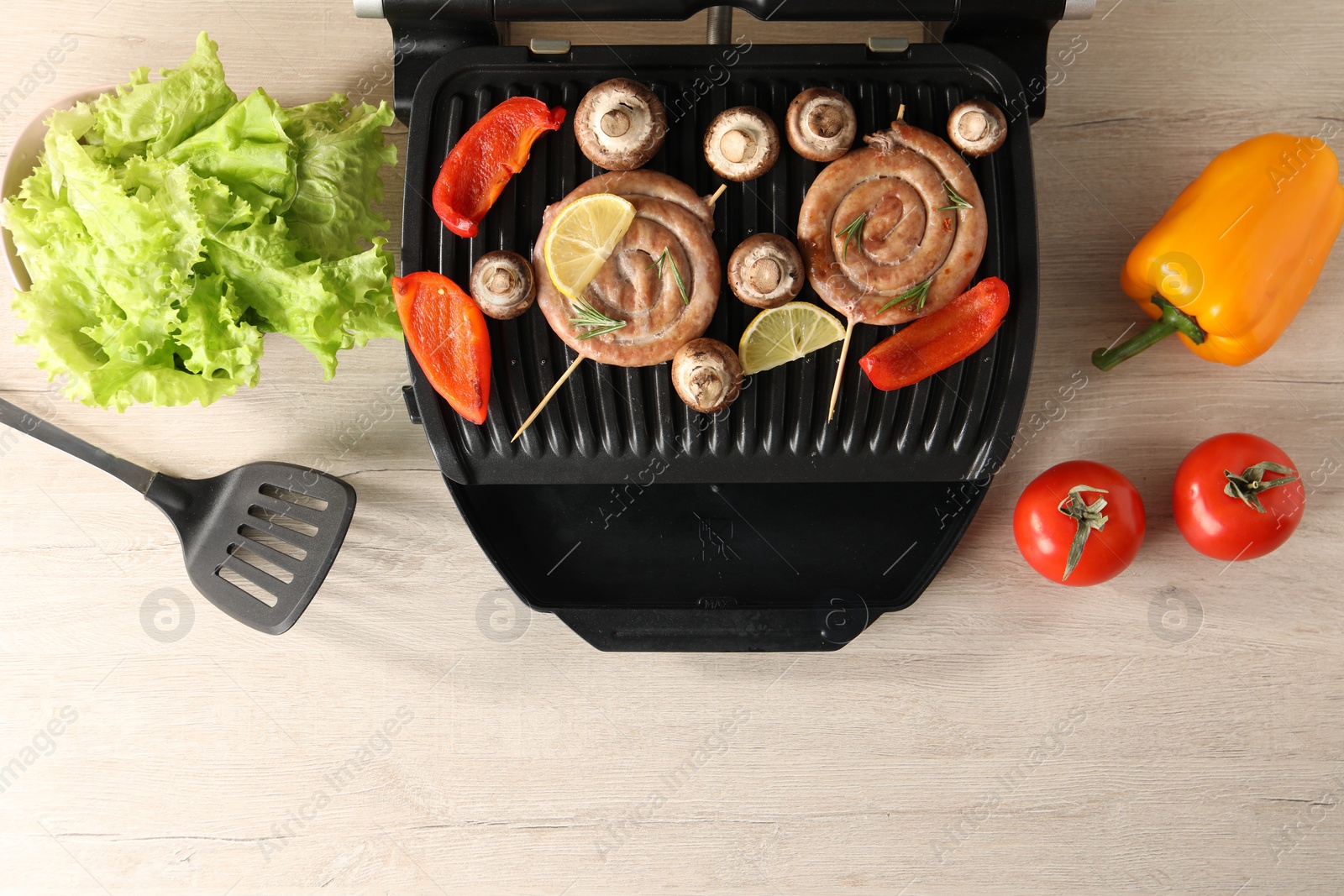 Photo of Electric grill with homemade sausages, vegetables and spatula on wooden table, flat lay