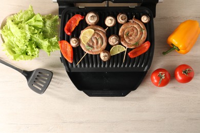 Photo of Electric grill with homemade sausages, vegetables and spatula on wooden table, flat lay