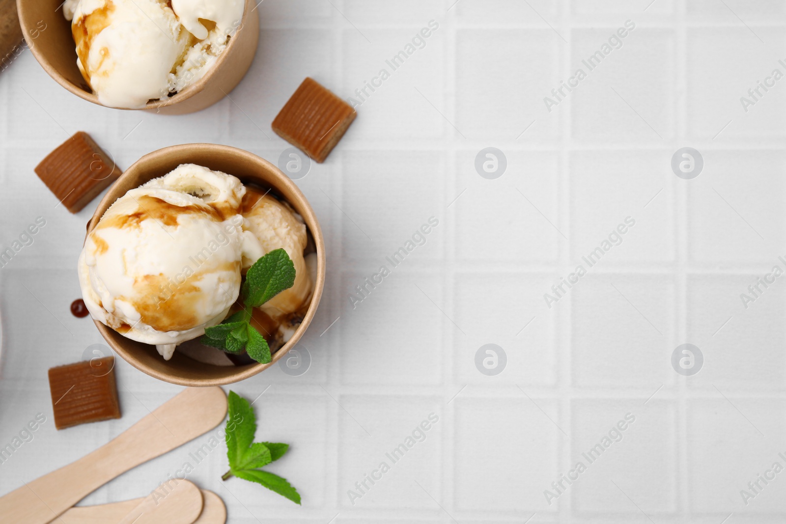 Photo of Scoops of ice cream with caramel sauce, mint leaves and candies on white tiled table, flat lay. Space for text