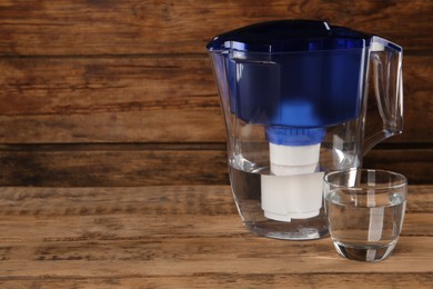 Photo of Filter jug and glass with purified water on wooden table. Space for text