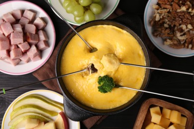 Photo of Dipping different products into fondue pot with melted cheese on black wooden table, flat lay