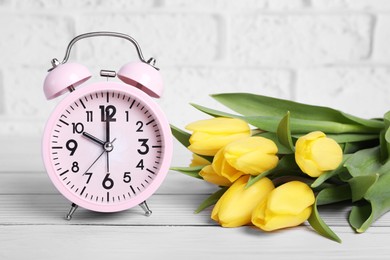 Photo of Pink alarm clock and beautiful tulips on white wooden table against brick wall. Spring time