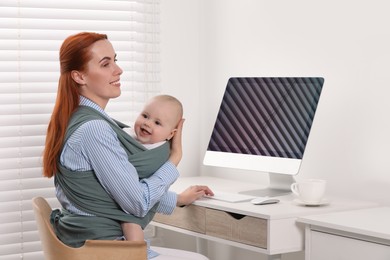 Mother holding her child in sling (baby carrier) while using computer at workplace