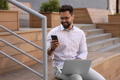 Photo of Handsome man using laptop and smartphone on bench outdoors