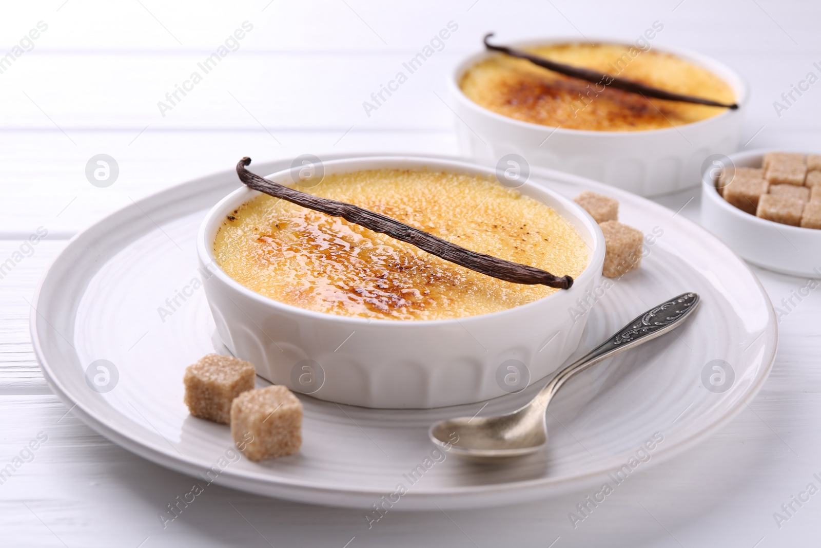 Photo of Delicious creme brulee in bowls, vanilla pods, sugar cubes and spoon on white wooden table, closeup