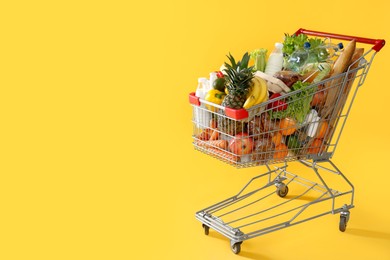 Photo of Shopping cart full of groceries on yellow background. Space for text
