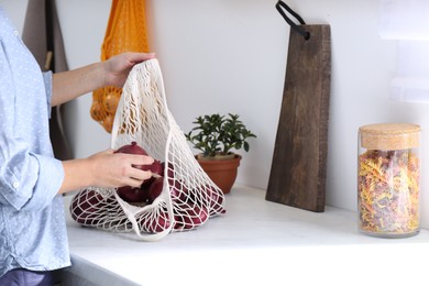 Photo of Woman taking red onion from mesh tote bag at countertop in kitchen, closeup