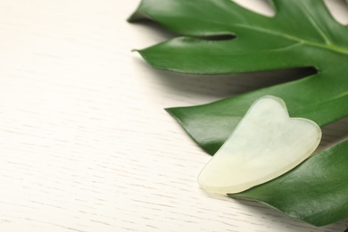 Jade gua sha tool and tropical leaf on white wooden table, space for text