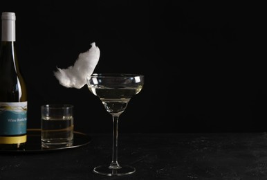 Photo of Cocktail with tasty cotton candy and bottle of alcohol drink on dark textured table against black background, space for text