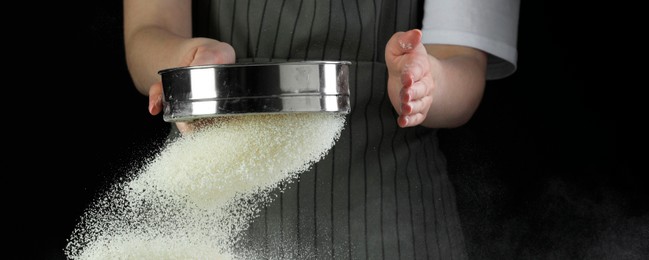 Photo of Woman sieving flour at table against black background, closeup